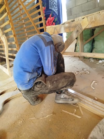 The scene depicts a woodworking workshop or construction area in Andijan, Uzbekistan. A single individual, appearing male, is captured from behind in a low squatting posture. The person wears a blue long-sleeved top with darker blue or black accents on the shoulders and arms, dark trousers heavily soiled with dust, and grey athletic shoes. Their hands, gloved in a light beige material, are raised to cover their face or head.
The ground is an earthen or concrete floor extensively covered with light-colored sawdust and wood shavings. Several pieces of unfinished wooden components, including curved sections and straight strips, are scattered on the floor near the individual. In the background, to the left, an upright wooden framework, possibly part of a furniture piece, is visible. To the right, a rough timber structure, potentially a workbench or support, is present. Behind this structure, a blue blanket featuring a prominent red floral pattern hangs, partially obscuring a bright natural light source. Further to the right, green bundled materials and rough grey concrete block walls are discernible. The environment suggests an active, dusty carpentry or construction work setting.