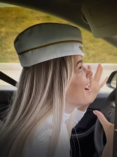 The image shows a close-up, profile view of a young woman wearing a white cap with gold trim, resembling a pilot's or flight attendant's hat. She is inside a vehicle, with her head turned to the right, her mouth open as if speaking or shouting, and her right hand raised towards her face. Her long, blonde hair cascades down her back and shoulders. The interior of the car is visible, including the steering wheel and part of the dashboard. The background outside the car appears to be a blurred, yellowish-green landscape, suggesting it might be a field or wooded area. The scene is likely captured during the daytime. There is no specific information within the image that would allow for its localization in Chorzów, Poland.