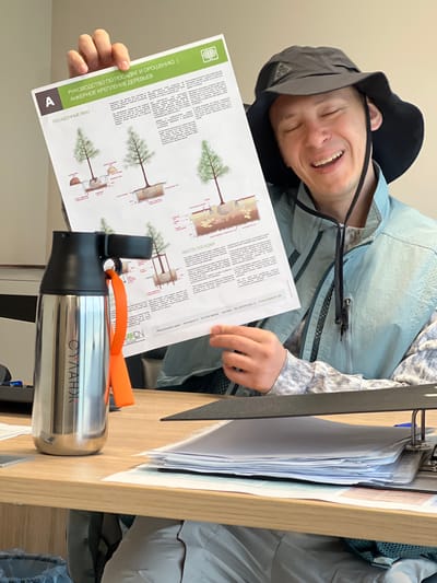 A person is seated at a desk, smiling and holding a document titled "РУКОВОДСТВО ПО ПОСАДКЕ И ОРОШЕНИЮ | АНКЕРНОЕ КРЕПЛЕНИЕ ДЕРЕВЬЕВ" (Guide to Planting and Irrigation | Anchor Fastening of Trees) and "ПОСАДОЧНЫЕ ЯМЫ" (Planting Pits) The document shows diagrams of tree planting methods. A stainless steel thermos with the name "СУЛАНЖ" is on the desk next to the document. The environment suggests an office setting. Based on the document, the location is potentially within a region where tree planting is relevant, such as Зеленогорское сельское поселение, Ukraine, because the document lists "Pflegepartnerschaften.Lappen.Hermannstal 14. D-41539 Meerbusch.Germany"