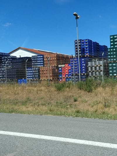 The image shows a fenced-off outdoor storage area with stacks of blue, red, brown, and green plastic crates, some containing bottles or kegs, under a clear blue sky. A white building with an orange roof is partially visible behind some of the stacks. A tall lamp post stands to the right of center. The foreground shows dry grass and a paved road with a white line. This scene is located in Barbing, Germany.