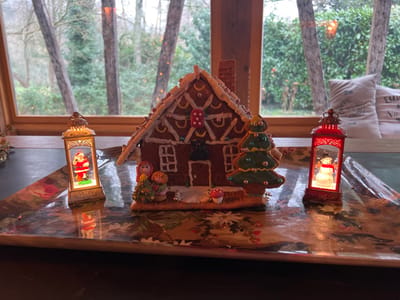 The scene depicts an indoor Christmas holiday display positioned on a reflective tray in front of a window, within an unspecified building in the city of Bailleul-la-Vallée, France.
At the center of the tray is a gingerbread house, detailed with white icing trim, a textured brown roof, and various confectionery elements including red and white circular candies. It features cut-out windows and a front door. To the left of the gingerbread house, a group of three small figurines, resembling a woman and two children, stands near a miniature green Christmas tree figurine adorned with colorful dots. A small red-capped mushroom figurine is also present on this side.
Flanking the gingerbread house are two illuminated lanterns. On the left, a vertical, ornate gold-colored lantern contains a figurine of Santa Claus dressed in a red suit, standing on a green base. On the right, a vertical red lantern contains a snowman figurine with a red scarf, positioned on a white, snowy base. Both lanterns emit internal light, creating reflections on the tray's patterned surface.
The background consists of a large, multi-paned window with a wooden frame, offering a view of an outdoor natural environment. Through the window, various trees, some with visible branches and others with green foliage, are discernible. Faint, warm lights, possibly decorative string lights, are visible in the distant greenery. To the far right, partially visible behind the right lantern, is a light-colored cushion or pillow, possibly made of burlap, with incomplete text visible on its surface. The overall lighting suggests an overcast day or early evening.