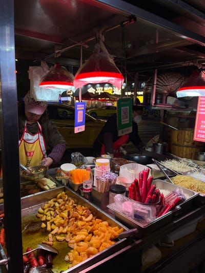 A street food vendor wearing a patterned hat and yellow apron stands behind a metal cart filled with various foods. There are containers of what appears to be fried potatoes, hot dogs, noodles, and dipping sauces. Red lamps with white trim are above the cart. Signs display Chinese characters, advertising the food. A QR code payment display is visible above the food preparation area. The scene is at night, under the glow of the streetlights.