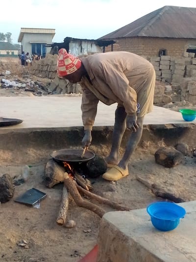 A man in a red and white patterned hat bends over a fire pit, stirring a pan with sticks. He wears a tan shirt, shorts, and yellow shoes. The fire burns with a small flame beneath the black pan. The scene takes place on a dusty, concrete-covered ground outside what appears to be some form of shelter. There are bowls nearby; one green and one blue. People and structures are visible in the background.