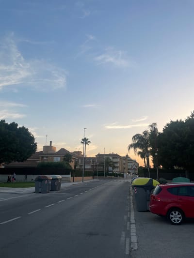 This is a vertical, low-angle outdoor photograph taken in Canet d'en Berenguer, Spain. The scene is a street with residential buildings and a clear sky. The time of day appears to be late afternoon or early evening, with a soft, warm light illuminating the sky and casting long shadows. The weather is clear, with only a few wispy clouds present.
In the foreground, on the right side of the image, a red car is parked alongside a large grey and yellow dumpster. A portion of the car's rear, including the wheel and taillight, is visible. Another dumpster is located further down the street. The street is paved and has white dashed lines indicating lanes.
The middle ground features a row of multi-story residential buildings with balconies. Palm trees and other lush green trees are scattered throughout the scene, adding to the Mediterranean feel. A street lamp with a single palm tree visible in front of it stands tall.
In the background, the sky transitions from a soft blue to a pale orange near the horizon, suggesting the setting sun. The overall mood of the image is peaceful and relaxed, conveying a sense of everyday life in a residential area. No text is clearly visible in the image.