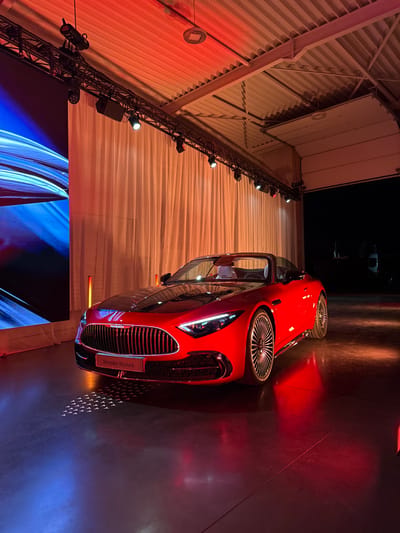 A red and black Mercedes-Maybach convertible luxury vehicle is centrally displayed indoors in Herentals, Belgium. The car is positioned at a slight angle, showcasing its prominent vertical-slat grille, sleek LED headlights, and multi-spoke wheels. Its interior features light-colored seating. The vehicle rests on a dark, reflective concrete floor illuminated by red and blue lights.

To the left, a large LED screen displays abstract blue and white light patterns, adjacent to two vertical, orange-glowing pillars. A long, light-colored curtain spans the mid-background. Overhead, a black metal truss system with spotlights is suspended from a corrugated metal ceiling, which is bathed in warm red-orange light. Small, triangular light patterns are projected onto the floor directly in front of the car. To the far right, a large industrial roll-up door is open, revealing a dark exterior with a white vehicle faintly visible in the distance. No individuals are present in the scene.