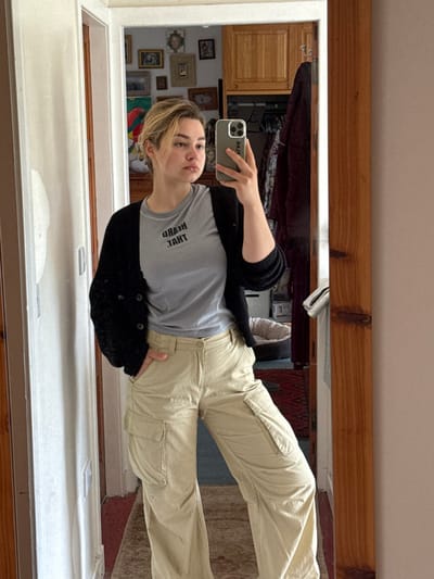 A young woman takes a mirror selfie. She wears a gray t-shirt with reversed text, a black cardigan, and light tan cargo pants. The photo is taken in a hallway with a wooden-framed mirror. Behind her, a closet door is open, revealing clothes and assorted items. Pictures and frames are visible on the wall next to the closet.