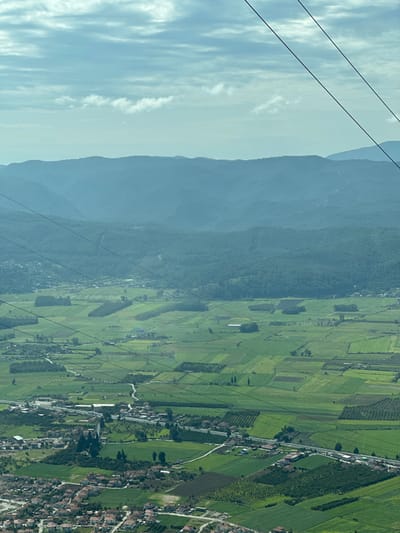 The image displays an elevated, wide-angle view of a rural and semi-urban landscape in Ula, Turkey, under a sky with scattered stratocumulus clouds against a light blue backdrop. In the furthest background, multiple layers of verdant, hazy mountains rise, their ridges undulating. Below these mountains, the mid-ground is dominated by an extensive plain of green agricultural fields, segmented into numerous distinct rectangular and irregular plots, some showing rows of cultivation, others appearing fallow or as pastures. Clusters of trees are interspersed within these fields, forming groves. A winding road traverses the fields, connecting various areas.

In the mid-foreground, a compact settlement, consistent with a town or village, is visible. This area contains numerous buildings, predominantly featuring light-colored walls and red-tiled roofs, characteristic of residential structures. A more prominent road or highway runs through and alongside this settlement, with vehicles occasionally visible as small dots. Utilities infrastructure is evident, with several dark, thin lines, consistent with power lines or support cables, prominently crisscrossing the entire frame from the upper left to the lower right, originating from an elevated vantage point outside the frame. No specific individuals or discernible actions are identifiable within the scene.