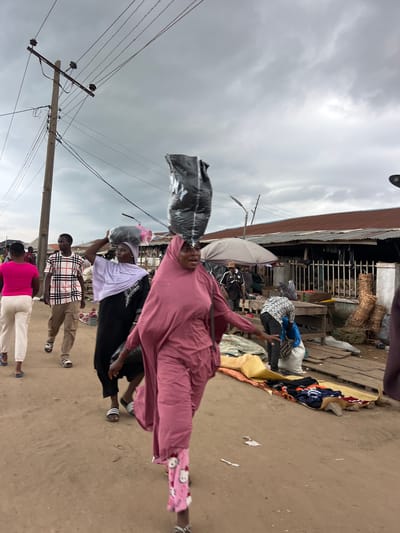 This is a medium-shot, eye-level, full-color, outdoor photograph taken at a market in Oko Amakom, Nigeria. The sky is overcast and gray, suggesting either a cloudy day or the onset of rain. A wooden utility pole with multiple insulators and wires is prominent on the left side of the frame, with some wires sagging. In the foreground, a woman wearing a rose-colored abaya and pink pants with white Hello Kitty patterns is walking towards the viewer, carrying a large black plastic bag on her head. She appears to be mid-stride, with her right arm extended forward. To her left, another woman wearing a black abaya and a lavender hijab is walking away from the viewer, also carrying a black plastic bag on her head. She is wearing white slides on her feet. A man in a white, pink, and black plaid shirt and beige cargo pants, wearing black and white slides, is walking behind her. Further left, a woman in a bright pink t-shirt and white pants is walking away from the viewer. In the background, a long, low building with a corrugated metal roof stretches across the frame. Several stalls with wooden tables and displays of goods, including woven baskets and what appear to be fabrics or clothing, are visible. Other individuals are present in the mid-ground and background, some appearing to be vendors or shoppers. The ground is dirt and appears dusty.