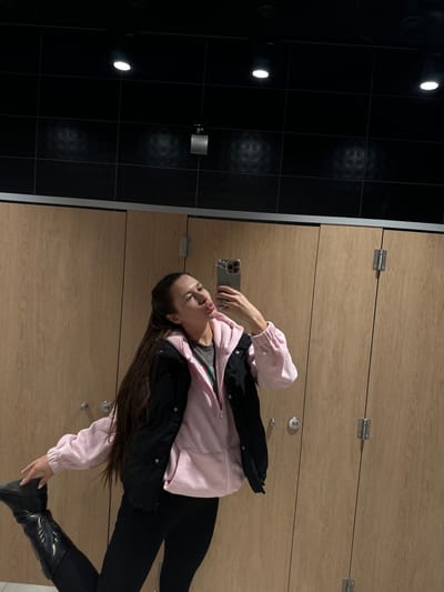A woman is taking a selfie in what appears to be a locker room. She is wearing black pants, a pink jacket over a black jacket and black boots. She is holding a phone in her hand. The room has wooden lockers and black tiled walls. There is no indication of the location being Ivashevo, Russia.