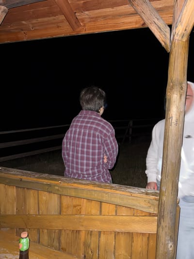 The image shows the interior of a wooden structure, possibly a shed or gazebo, at night. A person in a red and blue plaid shirt is visible within the structure. Another person wearing a white hoodie is partially visible above. A bottle can be seen in the lower right corner. The location is possibly Rila, Bulgaria, given the prompt's instructions.