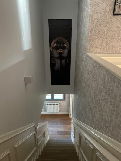 This image displays an interior residential or commercial stairwell and a lower-level hallway in Paracuellos De Jarama, Spain, viewed from an elevated perspective looking downwards. The carpeted staircase, in a neutral tone, is bordered by white wainscoting with decorative moldings on both sides. The left wall is painted a plain light grey, featuring a white rectangular wall sconce and a vertically oriented framed artwork. This artwork depicts a stylized dark-skinned individual, head tilted upwards with closed eyes, adorned with extensive gold-colored headwear and golden lips against a black background. The right wall is covered in light-colored wallpaper with a repeating botanical leaf pattern. The lower level, visible at the base of the stairs, features light brown wooden plank flooring and walls consistent with the botanical patterned wallpaper. This area contains a double window with white frames, beneath which a white panel radiator is affixed to the wall. A doorway is also visible on the right side of this lower space. Natural light illuminates the scene, with a vertical light reflection evident on the upper left wall. No individuals are present.