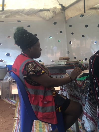 A person with dark skin sits on a blue plastic chair inside a structure covered in white plastic sheeting with circular holes. The person wears a red and gray striped safety vest over a patterned, multicolored shirt. The individual has black hair gathered on top of their head. At the top of the image, another person is partially visible, wearing a black top with "FILA" branding. A checkerboard pattern is also visible in their clothing. A table is nearby with various objects, including a container and a straw hat. The structure appears to be a temporary enclosure, as evidenced by the sheeting. The location is Nteje, Nigeria.