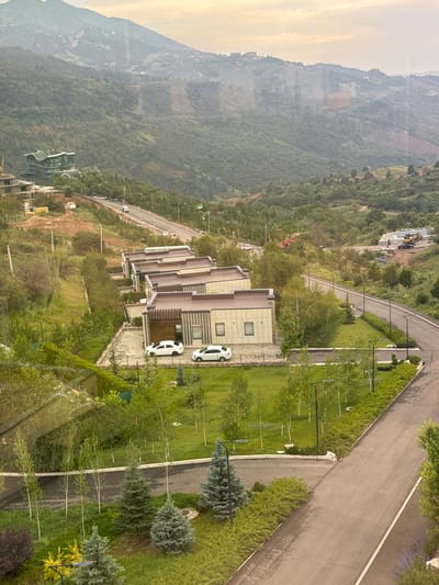 The image depicts a modern residential development nestled in a hilly, green landscape, likely on the outskirts of Uchterek, Uzbekistan. The foreground features a paved road winding through manicured lawns with meticulously planted trees and shrubs. Several contemporary, multi-unit dwellings with flat, brown roofs and beige facades are visible, each with a small yard and adjacent parking. Two white sedans are parked in front of one of the buildings. In the middle ground, the road continues uphill, with more construction activity visible in the distance, including excavators and portable buildings. Further up the slope, a larger, architecturally distinctive building with a curved, green roof and extensive glass windows is situated. The background is dominated by rolling, tree-covered hills and mountains, with a hazy sky. The scene appears to be an active construction and development area within a natural, semi-mountainous setting.