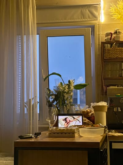 The image depicts a kitchen counter with a laptop displaying a woman in bed. To the left, sheer white curtains cover a window. A bouquet of white flowers in a vase sits on the counter, accompanied by glassware and bottles. In the background, a coffee machine is visible. The scene suggests a domestic setting, possibly a kitchen, with a focus on personal space and relaxation. The presence of the laptop and the woman's image implies digital engagement or entertainment. The overall ambiance is calm and intimate, with soft lighting and a hint of evening or dusk outside the window. The location, Тумілавіцкі сельскі Савет, Belarus, is not explicitly visible in the image but is provided as contextual information.