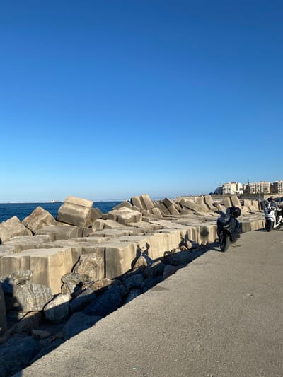 The image presents a view of a coastal area in Beau Fraisier, Algeria. Clear blue sky dominates the left side of the frame. A breakwater constructed of interlocking concrete blocks separates the sky from the sea. A paved road runs along the right edge of the frame. Two parked motorcycles are visible on the road, one black and one white. Buildings can be seen in the distance, situated above the breakwater. Shadows cast by the concrete blocks are present on the road.