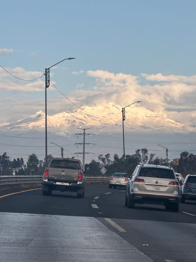 The image shows a daytime scene on a highway, presumably near Sanctorum, Mexico, based on the "PUEBLA" sign on a street lamp. Several vehicles are traveling on the road. In the background, a large mountain, possibly Popocatépetl, is visible, partially obscured by clouds. Power lines and vegetation are also present along the roadside. The sky is a mix of blue and clouds.