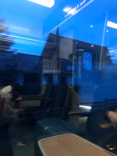 A blurry, skewed perspective within a train. Window reflections mingle with interior elements: a wall with small windows, seating, and overhead luggage racks. Passengers sit in the foreground, one with a phone. External light streams in, blurring the outside view, creating a layered, distorted composition.