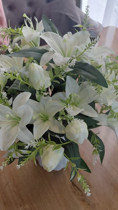 A close-up shot focuses on a bouquet of white artificial lilies and tulips arranged in a white vase. The flowers are diverse in bloom stage, with some fully open and others still in bud. The arrangement is adorned with artificial greenery, including long, slender leaves and delicate sprigs of small white flowers. The bouquet sits on a wooden surface, likely a table, which exhibits a natural wood grain pattern. In the blurred background, a hint of a person's shoulder and a glass containing a dark liquid can be discerned, suggesting an indoor setting. This scene is not specifically identifiable as being in Kerben, Kyrgyzstan, as no geographical markers are present in the image.