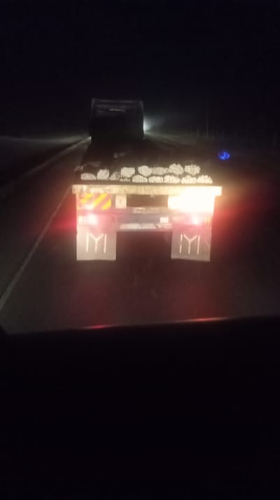 A nighttime shot from inside a vehicle, capturing the rear of a flatbed truck. The truck is loaded with cylindrical items. The taillights are illuminated, and each mud flap has a vertically oriented symbol. A vehicle is visible ahead in the distance. The scene suggests a road environment, potentially a highway, with limited visibility. The overall mood is dark and subdued.