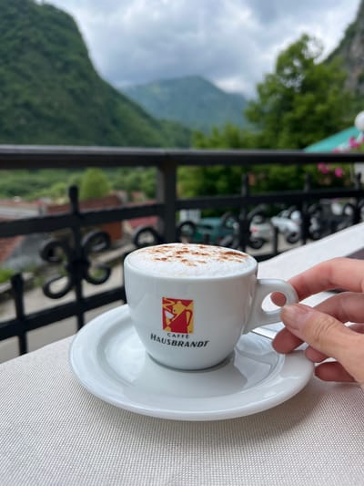 A close-up photograph depicts a hand gently holding the handle of a white ceramic coffee cup, resting on a matching saucer. The cup is placed on a light-colored, textured tablecloth. The beverage inside the cup features a creamy foam topping, lightly sprinkled with a brown powder, likely cinnamon or cocoa.
The side of the coffee cup prominently displays the brand name "CAFFÈ HAUSBRANDT" in black lettering, along with a distinctive red and yellow logo. This logo appears to be a stylized illustration of a figure, possibly a barista or a person enjoying coffee.
The setting is an outdoor area, likely a balcony or terrace, offering a scenic panoramic view. A black wrought-iron railing runs across the mid-ground, separating the table from the distant landscape. Beyond the railing, a vast, lush green mountainous terrain is visible under an overcast sky. Clouds are present, suggesting a cloudy or partly cloudy daytime. The image conveys a sense of tranquility, enjoying a coffee amidst a picturesque natural backdrop in Šavnik, Montenegro.