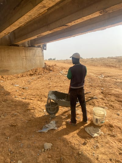 The image, when viewed in its correct orientation (rotated 90 degrees counter-clockwise), captures an outdoor scene featuring a person engaged in construction work under bright sunlight.
In the mid-ground, a person stands next to a metal wheelbarrow filled with what appears to be wet concrete or mortar. The individual is seen from behind and slightly to their left, wearing a light-colored baseball cap, a dark long-sleeved shirt with a hint of green fabric visible on their back, dark trousers, and athletic shoes. Their posture suggests they are either pausing from or preparing for work, possibly about to move the wheelbarrow.
The ground is dry, sandy, and light brown to reddish-brown, covered with small stones and scattered debris. Prominent, distinct shadows are cast by the person and the wheelbarrow, indicating direct and intense sunlight. To the right of the wheelbarrow, a white plastic bucket with some dark, illegible text on its side is visible, alongside a crumpled piece of white material, possibly a cloth or plastic bag.
On the left side of the frame, a substantial concrete structure spans horizontally across the view, appearing to be the underside of a bridge or an overpass. It consists of several thick, parallel concrete beams or girders, supported by a robust concrete pillar. The concrete shows signs of wear and has a rough, uneven texture in places, with some visible staining or rust.
The background on the right side of the image reveals more of the dry, elevated terrain, suggesting an undeveloped or excavated area. The sky, visible above the concrete structure and in the upper right, is bright and hazy, typical of a sunny day in a dry, dusty environment.
The scene depicts an active construction or maintenance site, likely involving the handling and transportation of concrete for the concrete structure. The time of day appears to be mid-morning to early afternoon, given the length and sharpness of the shadows. No clearly legible text is present in the image, other than the indistinct markings on the white bucket.