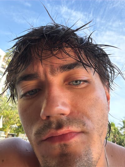 A close-up, upward-angle photograph shows a person with wet, dark hair and fair skin. The individual has light-colored eyes and stubble on their face. The background consists of a bright blue sky with light clouds and palm trees suggesting a coastal environment, possibly near Las Jarretaderas, Mexico. A building is partially visible in the background. A thin necklace is visible on the person's neck. The person is looking directly into the camera.
