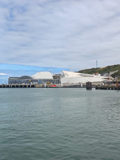 The image depicts a body of water in front of a modern, architecturally striking building complex in Boulogne-sur-Mer, France. The building has a prominent white, undulating roof that appears to be made of a lightweight material, possibly resembling a sail or a wave. To the left of this main structure, there is another building with a dark, curved roof and extensive glass facades. The complex is situated on the waterfront, with a pier or dock extending into the water. A small orange boat with white lettering, possibly a rescue or utility vessel, is moored alongside the dock. In the background, a green, tree-covered hill rises, topped with several buildings and a tall, slender antenna or tower. The sky above is partly cloudy with patches of blue. The water in the foreground is choppy, reflecting the sky and surrounding structures. There are no discernible people or specific actions taking place in the immediate foreground. The scene appears to be a daytime view of a coastal urban landscape, likely a port or marina area.