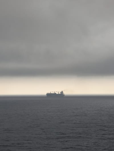 An overcast sky looms above a vast, dark ocean. In the distance, a cargo ship sails across the horizon. The ship is a long, rectangular vessel with cranes on its deck. A plume of smoke rises from the ship's stack, dissipating into the hazy atmosphere. The ocean is a deep, muted blue-gray, with small waves gently lapping against the hull of the ship. The overall scene evokes a sense of isolation and the immense power of nature. The image appears to be taken from a high vantage point, possibly a cliff or another ship, looking out over the water.