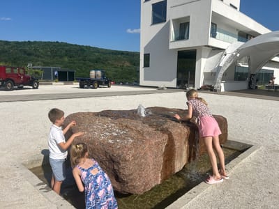In the city of Ulmu, Moldova, on a bright sunny day, three children are interacting with a water fountain situated in a courtyard. The fountain is constructed from a large, rough-hewn reddish-brown boulder with water spouting from its top and cascading down its sides into a surrounding concrete basin. A young boy, wearing a white polo shirt and light blue shorts, stands with his feet in the basin, reaching out towards the boulder. Beside him, a younger girl in a blue floral dress is also looking into the basin. To the right, a girl in a floral-print top and pink shorts leans over the boulder, her hands resting on its surface.
In the background, a modern white building with multiple levels and large windows is visible. To the left of the building, an arched white canopy structure is erected. Further left, a vintage red car is parked next to a dark grey building with a blue parking sign. Behind these structures, a green, forested hill rises under a clear blue sky. A flatbed truck carrying two wooden barrels is parked on the gravelly ground between the vintage car and the main building. The ground is covered in light-colored gravel.