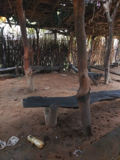 The image depicts an outdoor shelter constructed with natural materials. The roof appears to be thatched, supported by rough-hewn wooden posts and branches. A woven or stick-like barrier forms the background. The ground is covered in a sandy, earthy material, with some scattered debris including a plastic bottle and plastic bags. A dark, flat piece of wood, possibly a bench or table, is prominent in the foreground. The overall scene suggests a rustic, possibly rural, setting. The location is identified as Barikin Sibre, Nigeria.