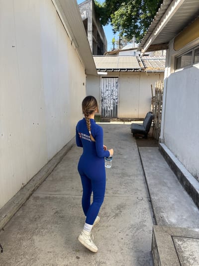 A woman wearing a blue jumpsuit stands outdoors holding a water bottle. The setting appears to be a narrow alleyway between buildings. Her hair is braided, and she wears white sneakers. The buildings are primarily white and appear to be in a state of slight disrepair. A gray concrete walkway stretches along one side. A few discarded items are visible in the background, adding to the somewhat industrial feel of the location.