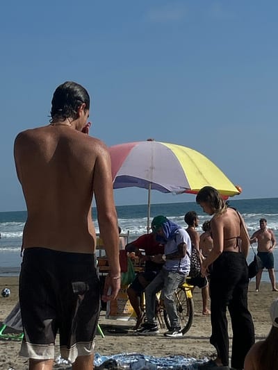 The image shows a beach scene in Montañita, Ecuador, located at coordinates -1.825 latitude and -80.754 longitude. A shirtless man with dark hair and a black bathing suit is standing on the beach with his back to the camera. A multi-colored beach umbrella is in front of him. The man is looking towards a woman standing next to a beach cart with a man in a green hat sitting on it. The woman is wearing a black bikini top and black pants. Other people are standing and walking on the beach, while the ocean with waves can be seen in the background. The beach is sandy and there are some blue and white blankets on the sand.