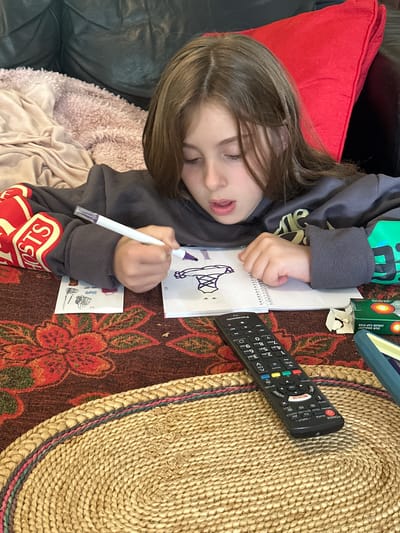 The image depicts a young girl with brown hair, wearing a dark grey hoodie, engaged in drawing on a notebook. She is holding a white marker with a purple tip and appears focused on her artwork. The notebook is open on a table with a floral patterned tablecloth. To the left of the notebook, there are several small sticker-like images. A black TV remote control is positioned in the foreground, near the center of the image. The girl is seated on a couch with a black leather back and a large red pillow. In the background, a light brown, fluffy blanket is visible. The scene appears to be indoors, possibly a living room. There is no information in the image that could be used to determine its location in the city of Thora, Australia.