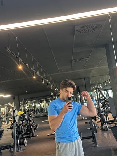 A fair-skinned man with brown hair is taking a selfie in a mirror at a gym. He is wearing a light blue t-shirt and gray sweatpants. He is holding a smartphone in his left hand and flexing his right bicep. Gym equipment is visible in the foreground and background, including weight racks, benches, and machines. The gym has black walls with vents, and there are track lighting fixtures along the ceiling. The photograph was taken in Jestetten, Germany.