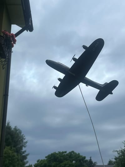 The image shows a silhouetted model airplane suspended in the air against a cloudy sky. The airplane is dark and appears to be made of plastic or a similar material. It has two propellers on its wings and a tail fin. A thin wire runs from the airplane to the right of the frame, suggesting it is being flown or displayed. The airplane has the registration "LY-AAA" visible on its fuselage. Below the airplane, a portion of a roofline and some greenery are visible, hinting at an outdoor setting. The presence of a roof and trees suggests this might be a residential area or a park. The sky is overcast, indicating a cloudy day, possibly with some diffused sunlight breaking through. No people are visible in the frame. The overall impression is of a whimsical display, possibly a toy or a decorative item. The location context indicates Lapės, Lithuania.