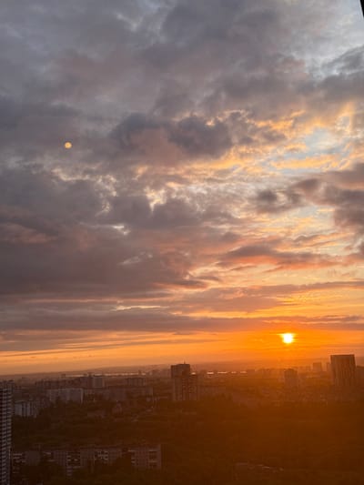 An urban landscape is depicted at sunset, viewed from an elevated vantage point. The sky occupies the majority of the frame, characterized by dense cloud cover transitioning in color. The upper-left quadrant features dark grey and bluish-grey clouds. Towards the horizon on the right, clouds are illuminated in shades of orange, yellow, and pink. The setting sun is visible as an intense yellow-orange disc on the right side, just above the urban horizon, partially obscured by atmospheric haze. A distinct, bright, textured circular artifact or reflection is present in the upper-left portion of the sky, against darker clouds. Below the sky, the city of Boldino, Russia, extends across the lower half of the image. It comprises a dense arrangement of multi-story buildings, including numerous residential apartment blocks and several taller high-rise structures, largely silhouetted against the bright sky. Patches of dark green vegetation are interspersed throughout the urban fabric, particularly in the mid-ground. A hazy distant urban area and a body of water are discernible on the far horizon. The foreground and mid-ground buildings are predominantly cast in shadow, while elements closer to the setting sun exhibit a warm, golden-orange hue. No people or specific actions are visible.