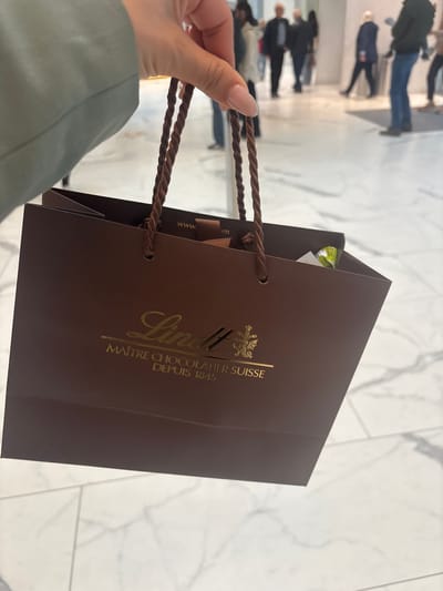 A person holds a dark brown Lindt chocolate shopping bag with a gold Lindt logo in what appears to be a well-lit shopping mall. The background shows multiple people walking on a marble-like floor. The image potentially portrays the aftermath of a purchase from a Lindt store, with the bag likely containing chocolates. Given Lindt's headquarters in Kilchberg (ZH), Switzerland, the shopping mall could be located in that area.