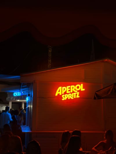 This image depicts a nighttime scene outside a building with a neon sign that reads "APEROL SPRITZ" in bright orange. The building appears to be a small establishment, possibly a bar or cafe, with horizontal wooden siding. To the left, a blue neon sign reads "COMIDA". Several people are visible in the foreground and background. Some are gathered near the building entrance, while others are seated at tables in the lower part of the frame, silhouetted against the ambient light. The overall atmosphere suggests a lively evening setting, likely in a coastal town given the location context of Xàbia/Jávea, Spain, known for its vibrant nightlife.