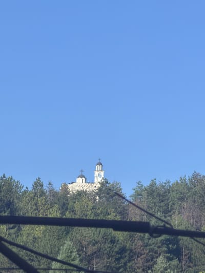 The image shows a clear blue sky above a dense forest. A building with a dome and a clock tower is visible on a hill, partially obscured by trees. In the foreground, a dark, horizontal structure, possibly a cable or a railing, runs across the image, out of focus. The building appears to be a church, possibly the Church of St. George in Hadzhidimovo, Bulgaria, given its architectural style and the context of the image. The forest is a mix of coniferous and deciduous trees. The scene is static, with no people or animals visible. The lighting suggests it is daytime.