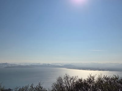 The image presents a wide-angle panoramic view of a serene body of water, likely a bay or sea, under a clear, bright sky with the sun visible as a lens flare in the upper right quadrant. In the distance, a range of mountains, capped with snow, forms a majestic backdrop. The water's surface is rippled, reflecting the sunlight in a shimmering pattern. In the foreground, the silhouettes of bare-branched trees and shrubs are visible, framing the expansive landscape. A small ship or boat is discernible on the water, and another larger vessel appears near the lower left edge of the frame. The scene suggests a natural, possibly coastal environment.