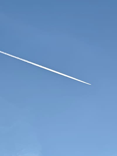 A daytime, outdoor shot depicts a clear, light blue sky with a linear contrail spanning the frame diagonally from the upper left to the lower right. The contrail is bright white, emanating from what appears to be an airplane at the lower right endpoint of the trail. The contrail dissipates slightly as it extends to the left. No land features or other objects are visible. The image does not provide sufficient information to confirm its location as Aumetz, France.