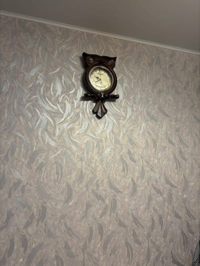 A close-up, high-angle shot captures a dark brown, ornate wall clock with a circular face and Roman numerals, mounted on a textured wallpaper. The wallpaper has a swirling pattern of light and dark grey brushstrokes, creating a dynamic and abstract design. The clock itself appears to be made of wood, with decorative carved elements resembling owl wings on either side of the face. The clock hands are visible, indicating a time between 8:00 and 9:00. A thin, white trim piece runs along the left edge of the frame, suggesting the clock is mounted on a wall in an indoor setting. The lighting is neutral, offering no indication of time of day or weather. No people are visible in the frame. The scene is static, with the clock being the sole focal point of the composition against the patterned background. The overall impression is one of decorative interior design.