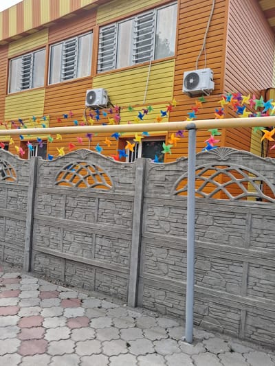 This image depicts the exterior of a building in Guldala, Kazakhstan. The building features a colorful facade with horizontal siding in shades of orange and yellow. Several windows with metal bars are visible on the upper floors. An air conditioning unit is mounted on the orange siding.
A decorative element consisting of a string of colorful pinwheels drapes across the front of the building, adding a playful and festive touch. Adjacent to the building is a gray concrete fence with a textured, stone-like pattern and decorative arched openings.
The ground is paved with interlocking concrete tiles in a light gray and pinkish-red pattern. A thin gray pipe runs horizontally across the frame, possibly part of the building's infrastructure. The scene appears to be outdoors during the daytime, with no specific indications of weather conditions. The overall impression is a bright and somewhat whimsical architectural setting. No people are visible in the image. There is no visible text.