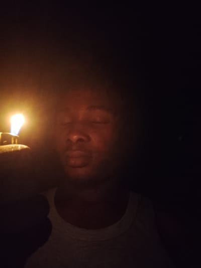 In a dimly lit environment in Orodo, Nigeria, a person is captured in a close-up shot, their face partially illuminated by the flame of a lighter held in their left hand. The person's eyes are closed, and their lips are slightly parted. They are wearing a light-colored tank top. The background is predominantly dark, with the intense light from the lighter creating a strong contrast and casting shadows. The overall atmosphere is intimate and perhaps contemplative, given the closed eyes and the solitary act of lighting the lighter.