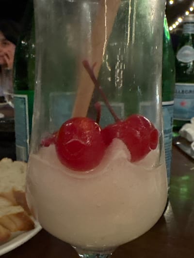 A close-up, slightly rotated, photograph depicts a tall glass containing a white, frothy beverage, garnished with two red cherries connected by a stem. A wooden swizzle stick is also visible inside the glass. Behind the glass, a green glass bottle of San Pellegrino sparkling water is partially visible. In the lower right corner of the frame, a white dish with what appears to be sliced bread can be seen. In the bottom left corner of the frame, part of a person's face can be discerned. This photograph was taken in Tamarindo, Costa Rica.