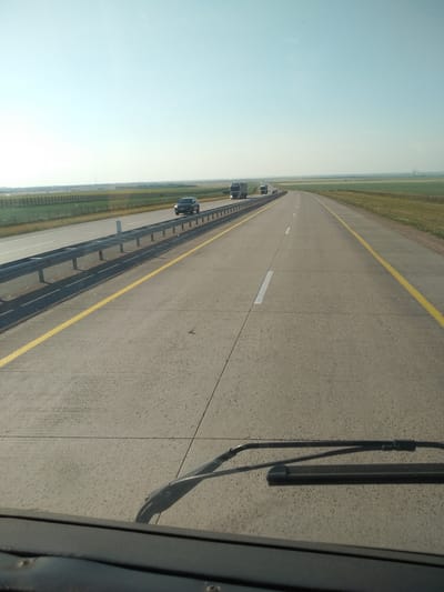 The image shows a clear, daytime view from inside a vehicle traveling on a highway. The road is paved with concrete and has yellow lane dividers. On the left side of the road, there is a metal guardrail separating the highway from a grassy field and a fence. Several vehicles, including trucks and a car, are visible on the highway ahead. The sky is clear and light blue, suggesting fair weather. The landscape on either side of the highway appears to be rural, with flat terrain and agricultural fields. The dashboard and windshield wipers of the vehicle are visible in the foreground, indicating the perspective is from the driver's seat. This scene is consistent with a highway in a rural area, and while Oskarovka, Kazakhstan has highways, no specific landmarks are visible to definitively place the location.