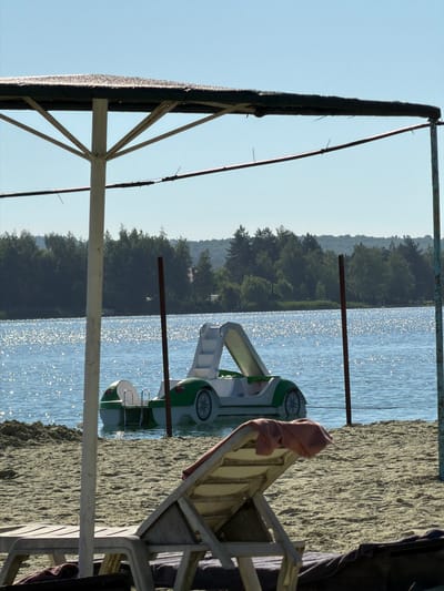 A sun-drenched beach scene in Verbizh, Ukraine, features a white and green pedal boat partially submerged in calm, glittering water. The boat, resembling a stylized car, has a white interior with steps leading up to a covered section. It is positioned in the middle distance, with a dense line of green trees forming the background across the water. In the foreground, a sandy beach is dotted with two empty lounge chairs. One chair, made of white plastic and wood, is in focus and has a pink towel draped over its back. A large, open beach umbrella with a dark mesh canopy and a white support pole dominates the left side of the frame, obscuring part of the sky and the lake. The sky is a clear, bright blue. The overall atmosphere is one of peaceful recreation.
