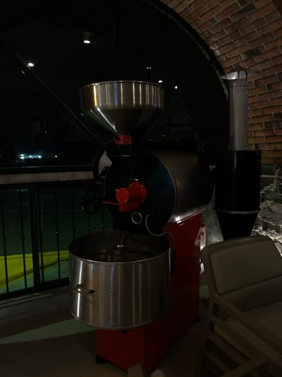 A coffee roasting machine is positioned indoors, presumably in Santa Ana Yancuitlalpan (Chilhuacán), Mexico, as specified. The machine features a large silver funnel on its left, leading into a black cylindrical central section. A red component is visible on this section. To the right, a larger silver cylinder is attached. A black exhaust pipe extends upwards, next to a chair with white cushions. The background shows a brick wall and stone masonry. Below the machine, a window with metal bars is visible, along with a yellow and green curtain. The floor appears to be tiled.