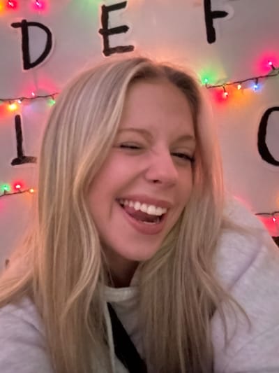 A smiling young woman with blonde hair, wearing a light-colored hoodie, is the main focus. Her face is slightly turned to the side, revealing her teeth. Behind her is a wall decorated with black letters arranged in a vertical row and a string of multi-colored Christmas lights. The overall image has a playful and informal feel, possibly a selfie or a candid shot.
