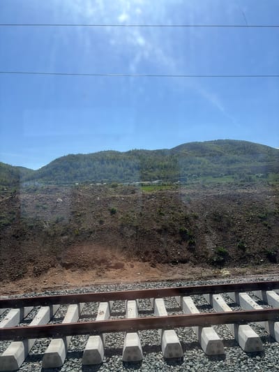 The image shows a view from a train window. On the left is a clear blue sky with power lines. In the center is a hillside covered with sparse vegetation and brown soil. On the right are railroad tracks with concrete sleepers and gravel. The location appears to be rural. The scene may be near Stambolovo, Bulgaria, given the prompt. No people or actions are visible.
